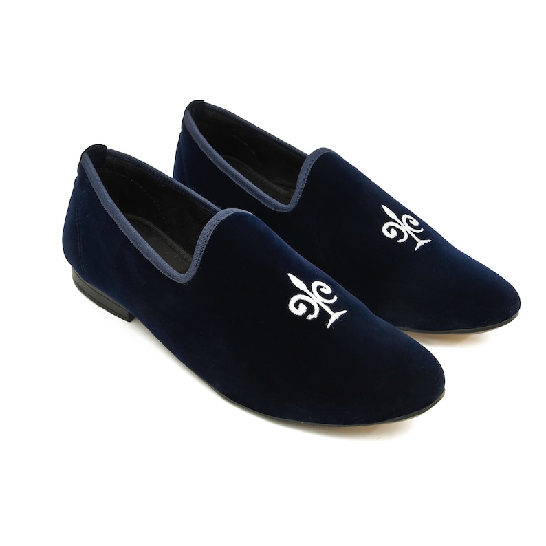 May include: Navy blue velvet loafers with white embroidered fleur-de-lis detailing. The shoes feature a dark blue trim and a low heel. These slip-on shoes are suitable for various occasions.