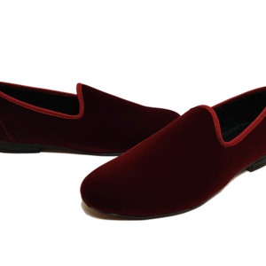 May include: Burgundy velvet loafers. These slip-on shoes have a rounded toe and a contrasting red trim around the opening. The soles are black. The shoes are a classic style.