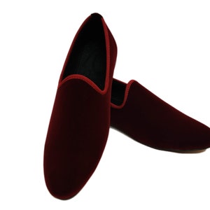 May include: A pair of burgundy velvet slip-on shoes. The shoes have a rounded toe and a red trim along the opening. The interior is black. The shoes are displayed on a white background.