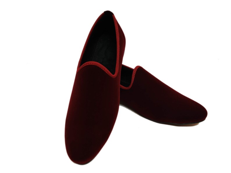 May include: A pair of burgundy velvet slip-on shoes. The shoes have a rounded toe and a red trim along the opening. The interior is black. The shoes are displayed on a white background.
