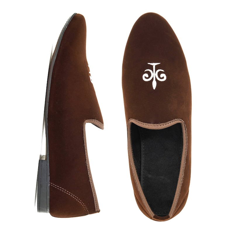 May include: A pair of brown velvet loafers with a white embroidered fleur-de-lis design.