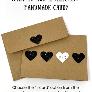 Op de afbeelding: Twee bruine enveloppen met zwarte glitterhartjes en een wit hartje met de tekst "10.6.18". De tekst "WANT TO ADD A MATCHING HANDMADE CARD?" staat bovenaan de afbeelding. De tekst "Choose the "+ card" option from the drop down menu when checking out" staat onderaan de afbeelding.