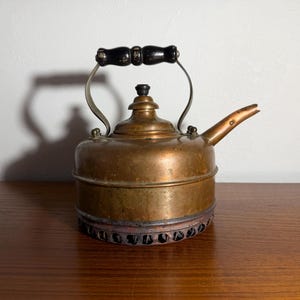 May include: Antique copper kettle with a black wooden handle and spout. The kettle has a round body and sits on a decorative base. The kettle is made of copper and has a vintage look.