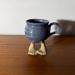 May include: A ceramic mug with a rounded, blue-glazed body featuring horizontal ridges and a single handle. The mug is supported by two sculpted, beige-colored feet with visible toes, giving the appearance that the vessel is standing upright.
