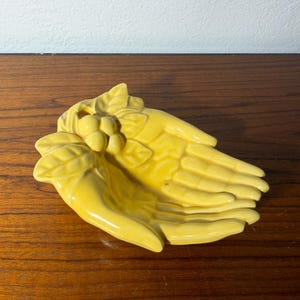 May include: A bright yellow ceramic trinket dish shaped like two cupped hands joined together. The design features a cluster of small, rounded berries and textured leaves at the wrist area, with a glossy finish covering the entire hand-shaped sculpture.