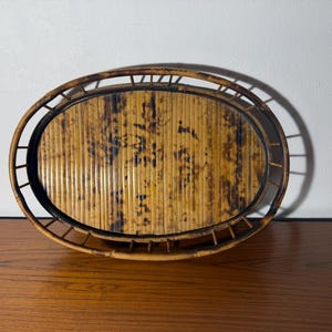 May include: An oval-shaped tray constructed from natural bamboo slats with a dark, mottled brown finish. The tray features a raised, slatted bamboo rim that encircles the central base, creating a distinct open-frame structure with a warm, earthy tone.