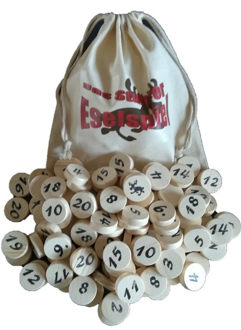 Puede incluir: Una bolsa de lona blanca con el texto "Das Spiel Eselspiel" en rojo y negro. La bolsa est&aacute; llena de c&iacute;rculos de madera con n&uacute;meros negros del 1 al 20.