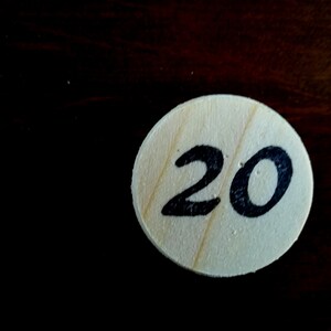 Puede incluir: Una ficha redonda de madera con el n&uacute;mero 20 escrito en tinta negra.
