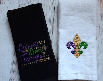 Könnte beinhalten: Ein schwarzes Handtuch bestickt mit dem violetten, grünen und goldenen Text 'Laissez les Bon Temps ROULER' und ein weißes Handtuch bestickt mit einem violetten, grünen und goldenen Fleur-de-Lis-Motiv. Beide Handtücher haben eine weiche Textur.