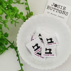 May include: White woven labels featuring a black sewing machine graphic and the text "I MADE THIS" in pink. The labels are in a white, scalloped bowl, with a sprig of green foliage. A white card with the text "JOSIE BUTTONS" is also visible.