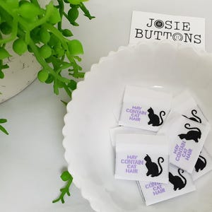 May include: White fabric labels with the text "MAY CONTAIN CAT HAIR" and a black cat silhouette. The labels are in a white scalloped bowl, with a "JOSIE BUTTONS" tag. Green plant in the background.