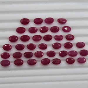 May include: A collection of thirty-three loose, oval-cut rubies arranged in six horizontal rows. Each gemstone features a deep red color with a faceted surface that reflects light, creating a uniform appearance across the set.