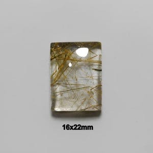 Puede incluir: Un cabujón de cuarzo rutilado rectangular que presenta un cuerpo claro y transparente con inclusiones doradas en forma de aguja. La piedra tiene una superficie lisa y pulida, y mide 16x22mm según el texto impreso debajo de la misma.