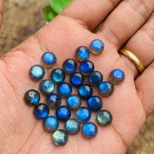 Puede incluir: Una colección de pequeñas piedras preciosas de labradorita pulidas y redondas con destellos azules iridiscentes. Cada piedra muestra una superficie de cabujón lisa y abovedada con varios tonos de azul profundo, verde azulado y gris.