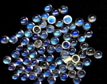 Puede incluir: Una colección de numerosas piedras de luna en cabujón, pequeñas, redondas y translúcidas. Cada gema presenta una superficie lisa y abovedada con una distintiva adularescencia azul que varía en intensidad sobre un fondo negro.