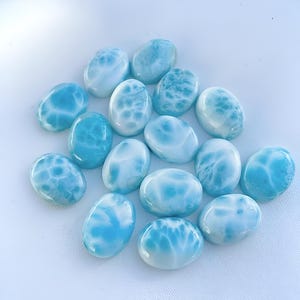 Puede incluir: Un conjunto de piedras preciosas de larimar ovaladas y pulidas. Las piedras varían en tonos azules y blancos, con patrones que recuerdan a las olas del océano. Las gemas están dispuestas sobre una superficie blanca, mostrando su belleza natural.
