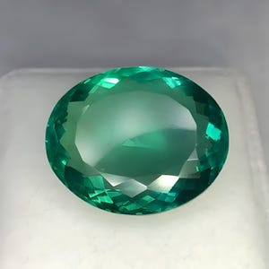 May include: A loose oval-cut gemstone with a vibrant teal-green color and a faceted surface. The stone displays high clarity and internal light reflection, showcasing a symmetrical arrangement of triangular and trapezoidal facets across its top.