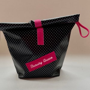 Pode incluir: Bolsa de maquiagem de bolinhas pretas e brancas com um zíper rosa e uma etiqueta de feltro rosa que diz "Beauty Queen".