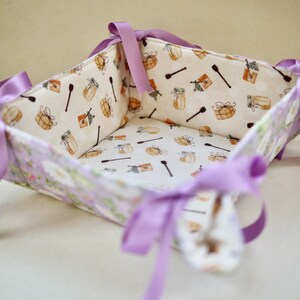 May include: A square fabric basket with a floral pattern on the outside and a food-themed pattern on the inside. The basket is trimmed with lilac ribbon tied into bows at each corner. The interior fabric features jars, honey dippers, and packages.