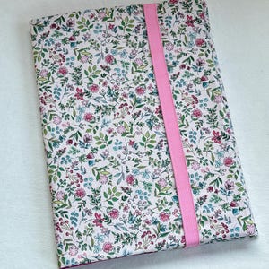 Puede incluir: Una funda para cuaderno con diseño floral y cierre elástico rosa. La funda presenta un fondo blanco con un patrón de flores y hojas rosas, azules y verdes. Una banda elástica rosa asegura la funda verticalmente.