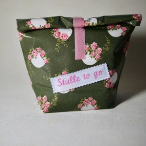 May include: A reusable lunch bag with a floral pattern of pink roses and white accents on a dark green background. The bag is secured with a pink velcro strap and features the embroidered text "Stulle to go".