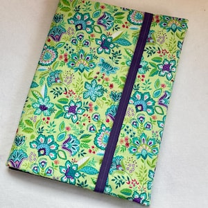Pode incluir: Uma capa de livro de tecido com um padrão floral e uma faixa elástica roxa. A capa apresenta um fundo verde claro com um padrão repetido de flores e folhas turquesas, roxas e vermelhas. A capa é retangular.