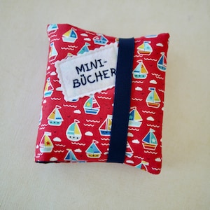 Könnte beinhalten: Eine quadratische Stofftasche mit rotem Hintergrund und einem wiederkehrenden Muster aus kleinen Segelbooten in Blau, Gelb und Weiß. Ein dunkelblaues Gummiband umschließt die Mitte und ein weißer Filzaufnäher mit dem Text MINI-BÜCHER ist angebracht.