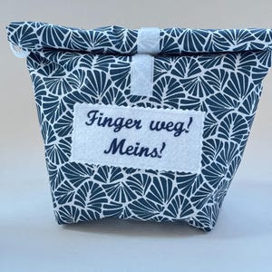 May include: A white fabric bag with a blue and white geometric pattern. The bag has a velcro closure and a white label with the text "Finger weg! Meins!" embroidered in blue.