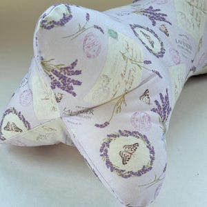 May include: A lavender-colored, bone-shaped pillow with a repeating pattern of lavender sprigs, butterflies, and vintage-style stamps. The pillow is made of soft fabric and is designed for comfort and support.