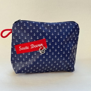 May include: Navy blue zippered pouch with a white anchor pattern. A red rectangular patch with the text "Seute Deern" is attached. The pouch has a red loop and a silver-colored charm.