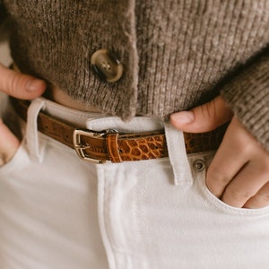 Peut inclure: Un cardigan en tricot marron avec quatre boutons et une ceinture en cuir marron avec une boucle dorée. Le cardigan est porté sur un jean blanc.