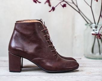 May include: A pair of brown leather ankle boots with lace-up fronts and block heels. The boots are on a white wooden surface.