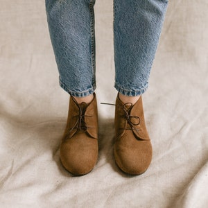 May include: A pair of brown suede ankle boots with laces. The boots are worn with blue jeans and a brown hat.