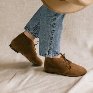 May include: A pair of brown suede ankle boots with laces. The boots are worn by a person wearing blue jeans and a brown hat.