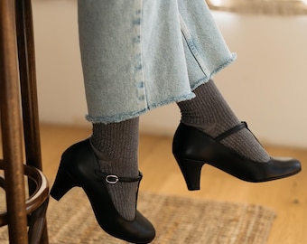 Puede incluir: Un par de zapatos negros de tacón con una hebilla. Los zapatos se llevan con calcetines grises y pantalones vaqueros azules.
