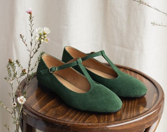 May include: A pair of dark green suede T-strap flats featuring a rounded toe, a thin ankle strap with a small gold-toned metal buckle, and a flat black sole. The interior of the shoes is lined with a light tan material, showing visible stitching.