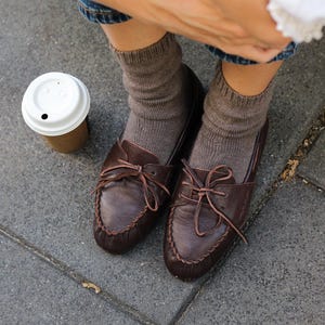 May include: A pair of dark brown leather moccasins with thin laces tied in bows at the top, worn with ribbed taupe-coloured socks. The shoes feature a stitched moc-toe construction and a smooth finish, paired with blue denim jeans and a white top.