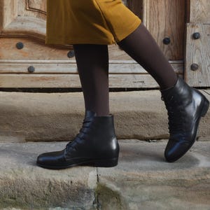 Puede incluir: Un par de botines de cuero negro con cordones, combinados con medias opacas de color marrón oscuro y una falda hasta la rodilla de color amarillo mostaza. Los botines tienen punta redondeada, tacón bajo y acabado liso, vistos de perfil.