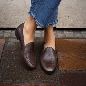Può includere: Mocassini in pelle marrone scuro. Queste scarpe slip-on hanno un design classico con punta arrotondata. L'immagine mostra le scarpe dal basso, con i piedi di una persona che indossa jeans blu.