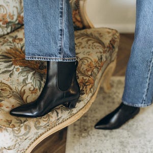 Puede incluir: Un par de botines de cuero negro con punta afilada, tac&oacute;n bajo y paneles el&aacute;sticos laterales, combinados con pantalones vaqueros azules. Los botines presentan un acabado liso y una trabilla en la parte posterior del tobillo para facilitar su uso.