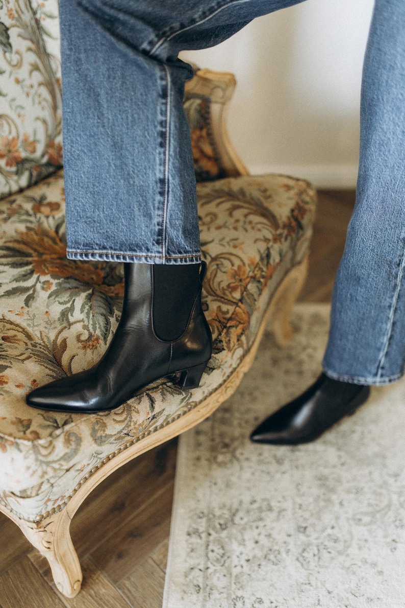 Puede incluir: Un par de botines de cuero negro con punta afilada, tac&oacute;n bajo y paneles el&aacute;sticos laterales, combinados con pantalones vaqueros azules. Los botines presentan un acabado liso y una trabilla en la parte posterior del tobillo para facilitar su uso.