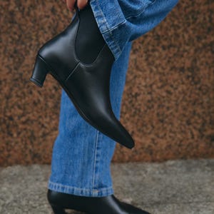 Puede incluir: Un par de botines Chelsea de cuero negro con punta afilada, tac&oacute;n bajo de bloque y paneles laterales el&aacute;sticos. Los botines se llevan con vaqueros azules, con uno de ellos levantado para mostrar el perfil lateral y el otro apoyado en el suelo.