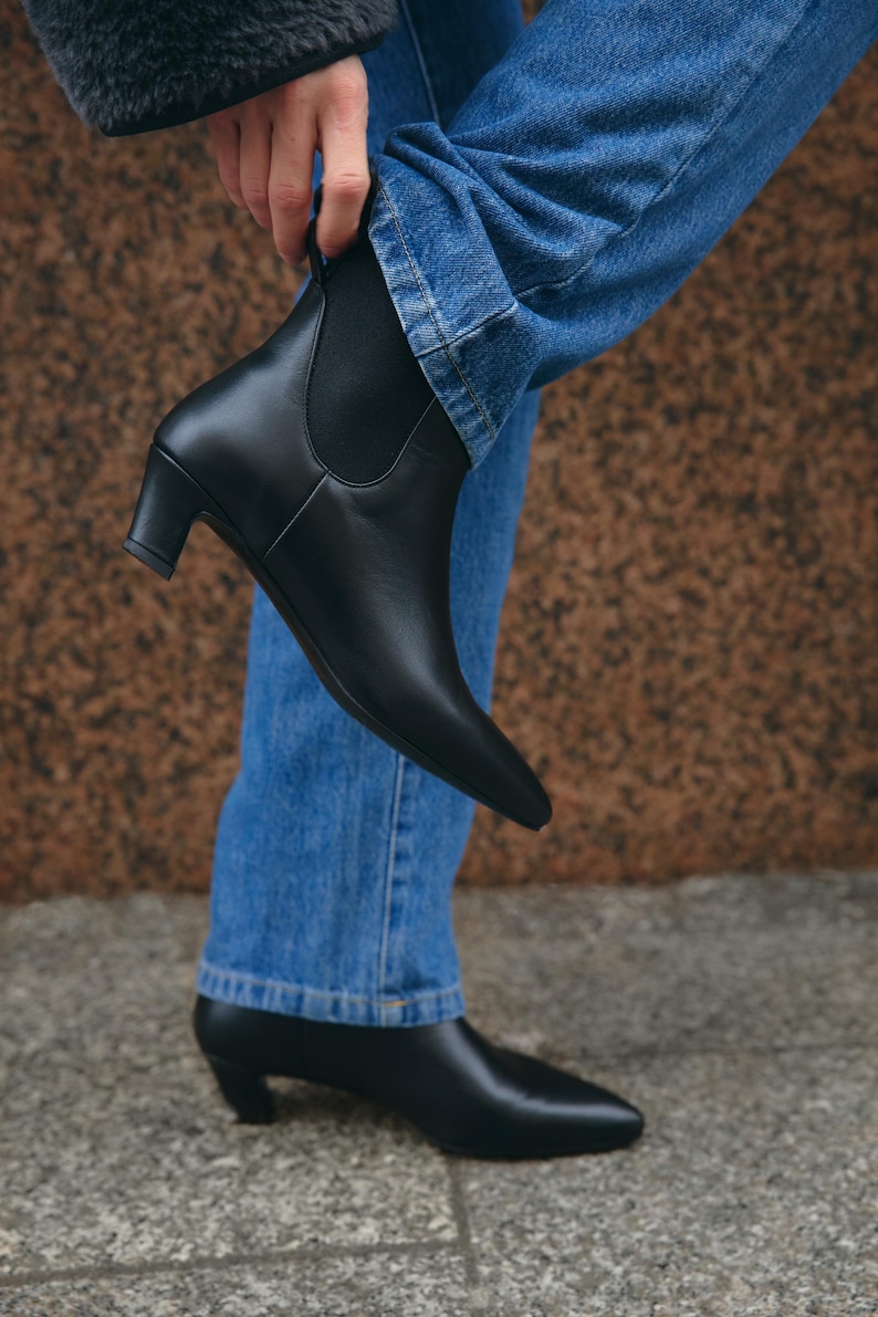 Puede incluir: Un par de botines Chelsea de cuero negro con punta afilada, tac&oacute;n bajo de bloque y paneles laterales el&aacute;sticos. Los botines se llevan con vaqueros azules, con uno de ellos levantado para mostrar el perfil lateral y el otro apoyado en el suelo.
