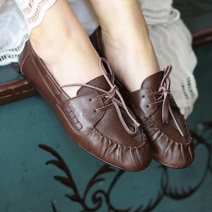Puede incluir: Un par de zapatos de cuero marr&oacute;n oscuro estilo mocas&iacute;n con costura fruncida en la punta, un panel lateral decorativo con detalles de costura y cordones finos de color claro atados en un lazo en la parte superior del empeine. Tienen un acabado liso.