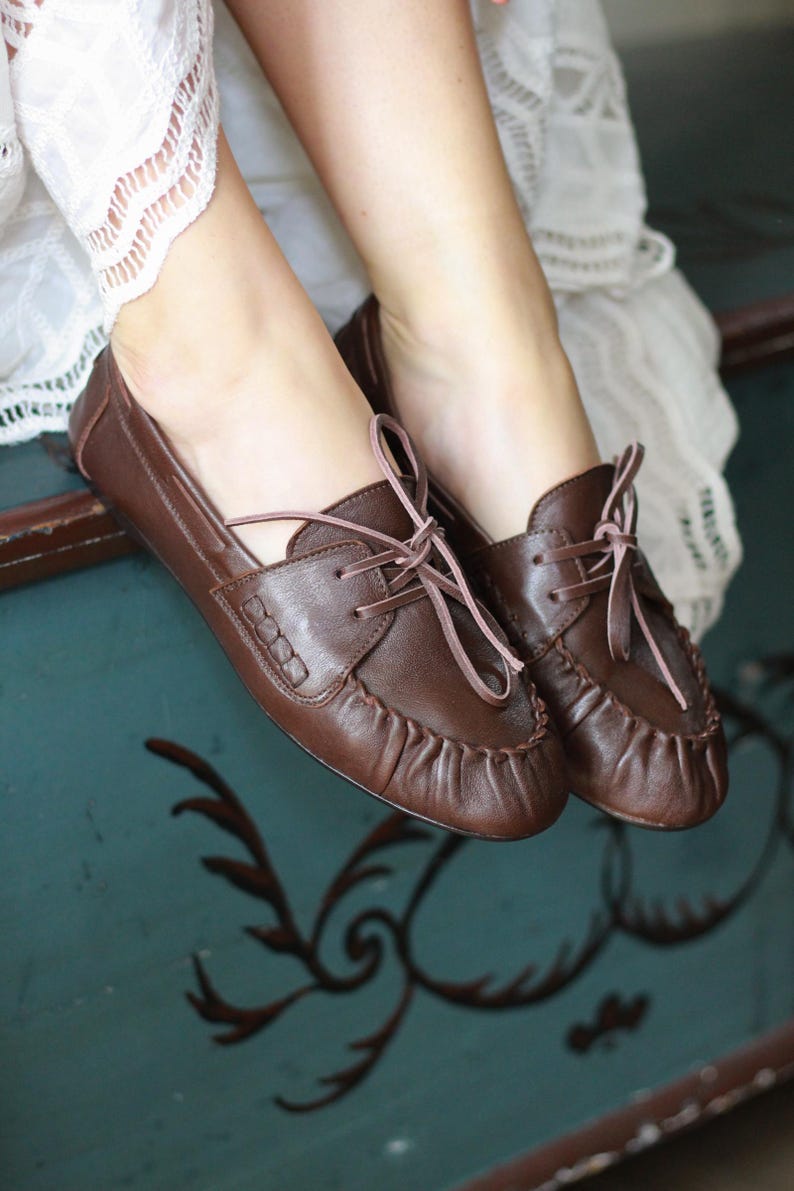 Puede incluir: Un par de zapatos de cuero marr&oacute;n oscuro estilo mocas&iacute;n con costura fruncida en la punta, un panel lateral decorativo con detalles de costura y cordones finos de color claro atados en un lazo en la parte superior del empeine. Tienen un acabado liso.