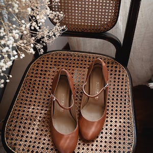 May include: A pair of brown leather heeled shoes with a buckle strap on a woven chair seat.