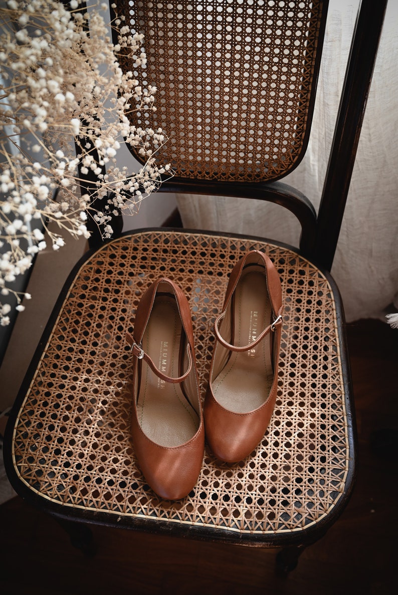 May include: A pair of brown leather heeled shoes with a buckle strap on a woven chair seat.