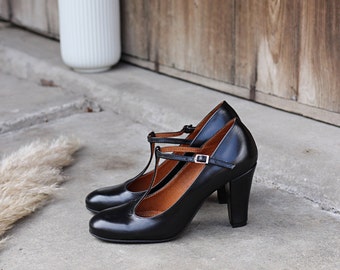 Puede incluir: Un par de zapatos de tacón negros con tira en T, con punta redondeada, tacón de bloque resistente y una tira fina ajustable en el tobillo con una pequeña hebilla de metal plateado. El forro interior de los zapatos es de color beige.