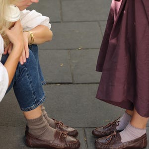 Puede incluir: Dos pares de zapatos de cuero marr&oacute;n oscuro estilo mocas&iacute;n con cordones finos y costuras visibles en la puntera. Un par se lleva con calcetines beige acanalados y el otro con calcetines tobilleros de color lavanda con borde rizado.