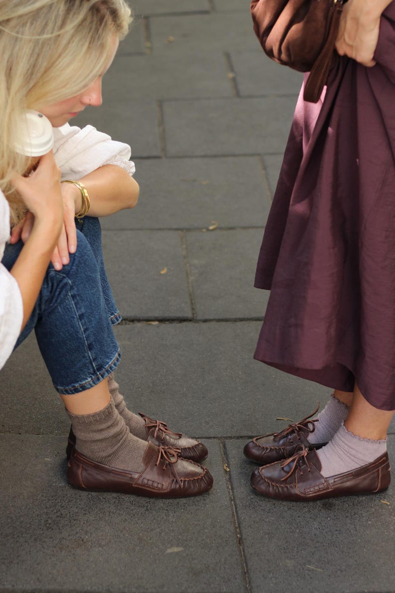 Puede incluir: Dos pares de zapatos de cuero marr&oacute;n oscuro estilo mocas&iacute;n con cordones finos y costuras visibles en la puntera. Un par se lleva con calcetines beige acanalados y el otro con calcetines tobilleros de color lavanda con borde rizado.